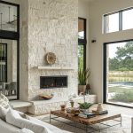 Luxury living room with fireplace, white stone wall, and black steel windows in modern style, with dining table near glass door to outdoor patio.